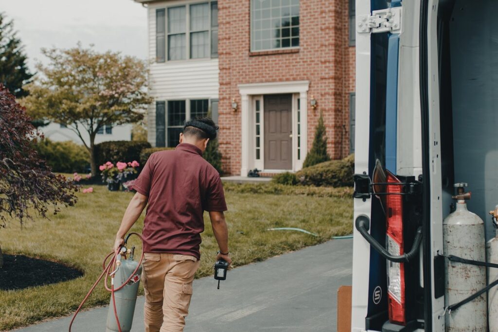 HVAC technician walking up a driveway toward a brick two-story home beside an open service van